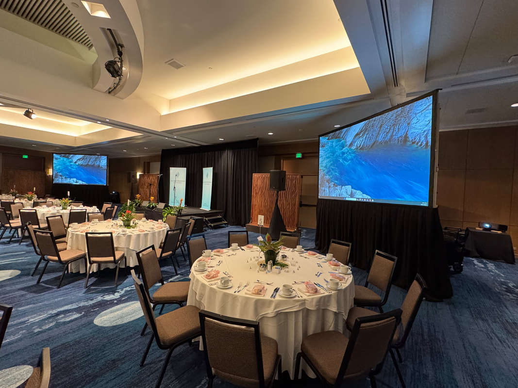 Two projection screens and a stage set up in a ballroom with round banquet tables