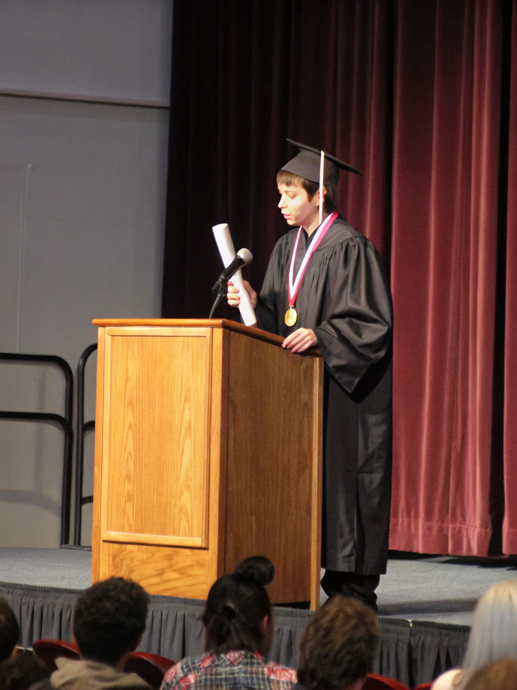 Graduate in cap and gown speaking at an indoor ceremony podium.
