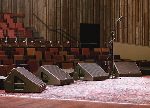 Four black stage monitor wedges on a wooden stage with microphone stands behind